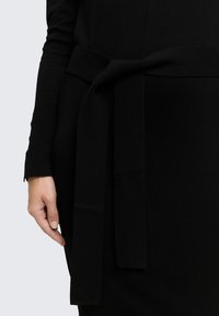 Mano reposando a un lado, vistiendo una prenda de manga larga negra con un cinturón de tela ancho atado en un nudo suelto en la cintura.