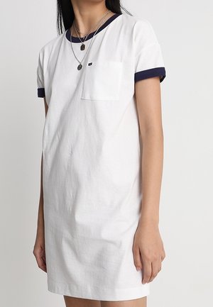 Biała sukienka z krótkim rękawem z granatowym wykończeniem przy rękawach i dekolcie, z kieszonką na piersi, noszona przez osobę noszącą warstwowe srebrne naszyjniki.