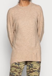 Beiger Strickpullover mit langen Ärmeln, geripptem Halsauschnitt und lockerer Passform, kombiniert mit grünen Camouflage-Musterhosen. Weicher Stoff.