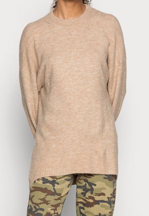 Beige stickad tröja med långa ärmar, ribbad halsringning och lös passform, tillsammans med byxor i grön kamouflage. Mjuk textur.