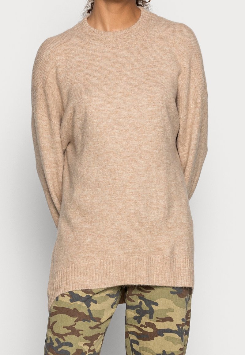 Beiger Strickpullover mit langen Ärmeln, geripptem Halsauschnitt und lockerer Passform, kombiniert mit grünen Camouflage-Musterhosen. Weicher Stoff.