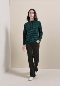 Zielony i czarny paskowany sweter z kapturem na sznurki, w połączeniu z czarnymi prostymi spodniami i białymi sneakersami. Gładkie tło.