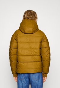 Padded jacket v hořčicově žluté barvě, s kapucí, horizontálním prošíváním a žebrovanými manžetami. Nosené s modrými džíny.