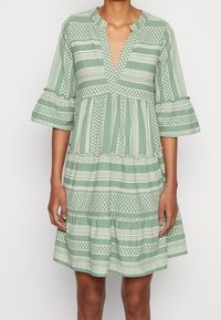 Grön klänning med korta ärmar och vita geometriska mönster, V-ringning, lager-på-lager design och volangärmar, gjord av lätt tyg.