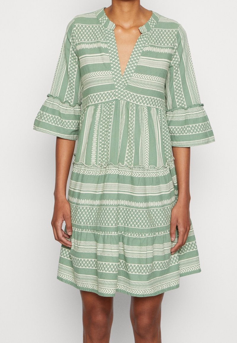Grön klänning med korta ärmar och vita geometriska mönster, V-ringning, lager-på-lager design och volangärmar, gjord av lätt tyg.