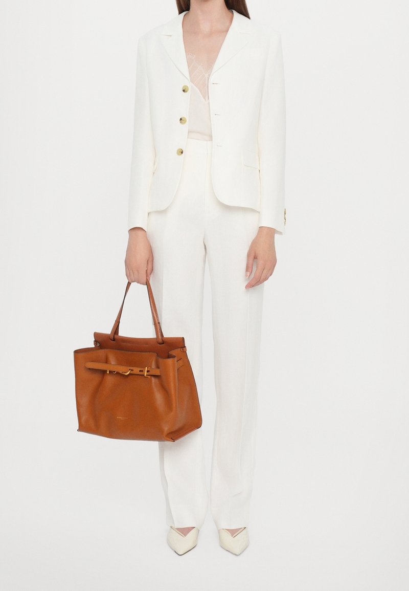 Femme en tailleur-pantalon blanc tenant un grand sac à main en cuir beige avec double poignée et détail de boucle.