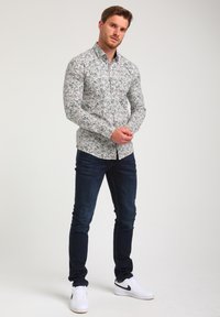 Langarm-Hemd aus leichtem Stoff mit einem bunten Blättermuster, kombiniert mit dunkelblauen Jeans und weißen Sneakers.