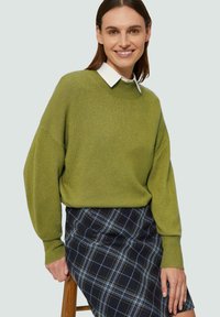 Grüner Strickpullover mit Rundhalsausschnitt und überschnittenen Schultern, kombiniert mit einem blauen Karorock, der weiße und gelbe Akzente aufweist.