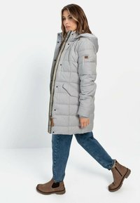 Grå pufferjakke med hætte, quiltet tekstur, forlommer og kontrasterende indvendig foring. Sammen med blå jeans og brune ankelstøvler.