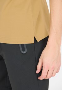 En tanfärgad t-shirt med kort ärm parat med svarta byxor med en sidoficka och strukturerad tyg, uppvisad mot en neutral bakgrund.