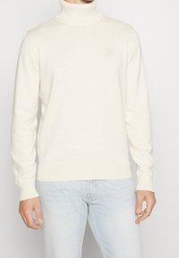 Man i en krämfärgad polotröja och ljusblå jeans, stående mot en enfärgad bakgrund.