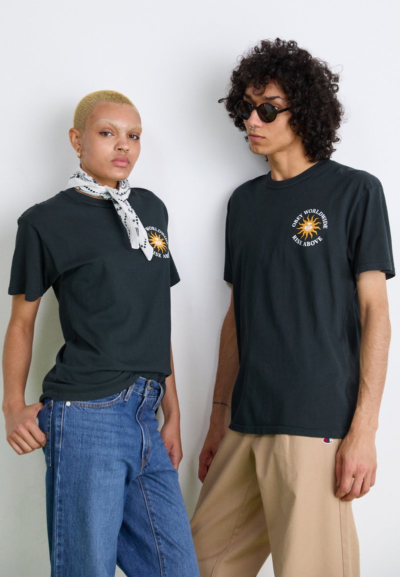 Svarte bomull t-skjorter med et grafisk solmotiv foran. Kvinnen har på seg et mønstrete hvitt skjerf og jeans; mannen har på seg beige bukser.