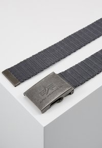 Ceinture en tissu tissé gris avec une boucle en métal portant un logo. La boucle a une finition usée ; la texture de la ceinture est tricotée serrée et durable.