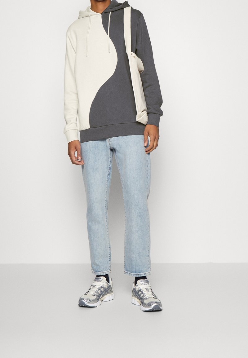 Personne portant un sweat à capuche bicolore noir et blanc, un jean bleu clair, des baskets argentées et un sac à bandoulière couleur crème.