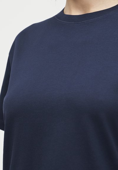 Κοντινό πλάνο ενός ατόμου που φοράει ένα απλό μπλε σκούρο T-shirt με λαιμόκοψη και κοντά μανίκια, σε λευκό φόντο.