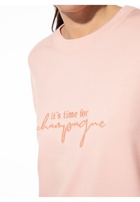 Lichtroze sweatshirt met een ronde hals, voorzien van oranje geborduurde tekst die zegt "het is tijd voor champagne." Zacht katoenen blendmateriaal.