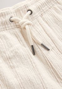 Primo piano di pantaloni in tessuto beige dalla trama strutturata con fascia elastica in vita e coulisse color crema annodata con punte in metallo.