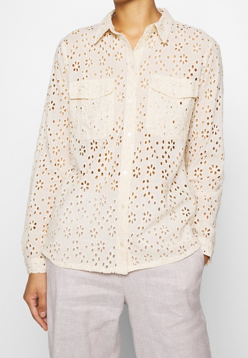 Camicia bianca, leggera, con bottoni, caratterizzata da un intricato motivo floreale ad occhiello, due tasche frontali e maniche lunghe con polsini abbottonati.