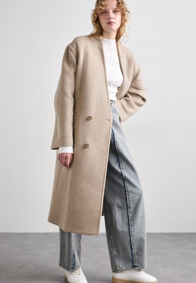 Beige uldfrakke med løs pasform, dobbeltknap-til design, store knapper og sidelommer, lagdelt over en hvid ribbet sweater og lyse jeans.