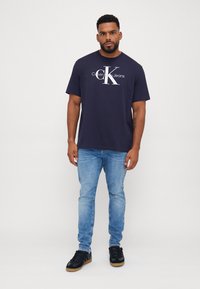 Camiseta de algodón azul marino con el logo en blanco de "Calvin Klein Jeans", combinada con jeans de denim azul claro y zapatillas negras con suelas de goma.