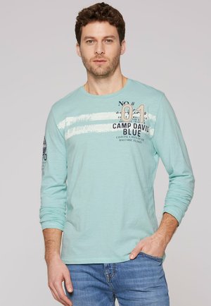 Uomo con capelli ricci che indossa una camicia azzurra a maniche lunghe con testo stampato e jeans blu su uno sfondo uniforme.