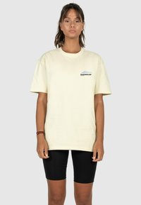Light yellow cotton t-shirt with a crew neck, short sleeves, and a small blue and black logo on the left chest, paired with black shorts.