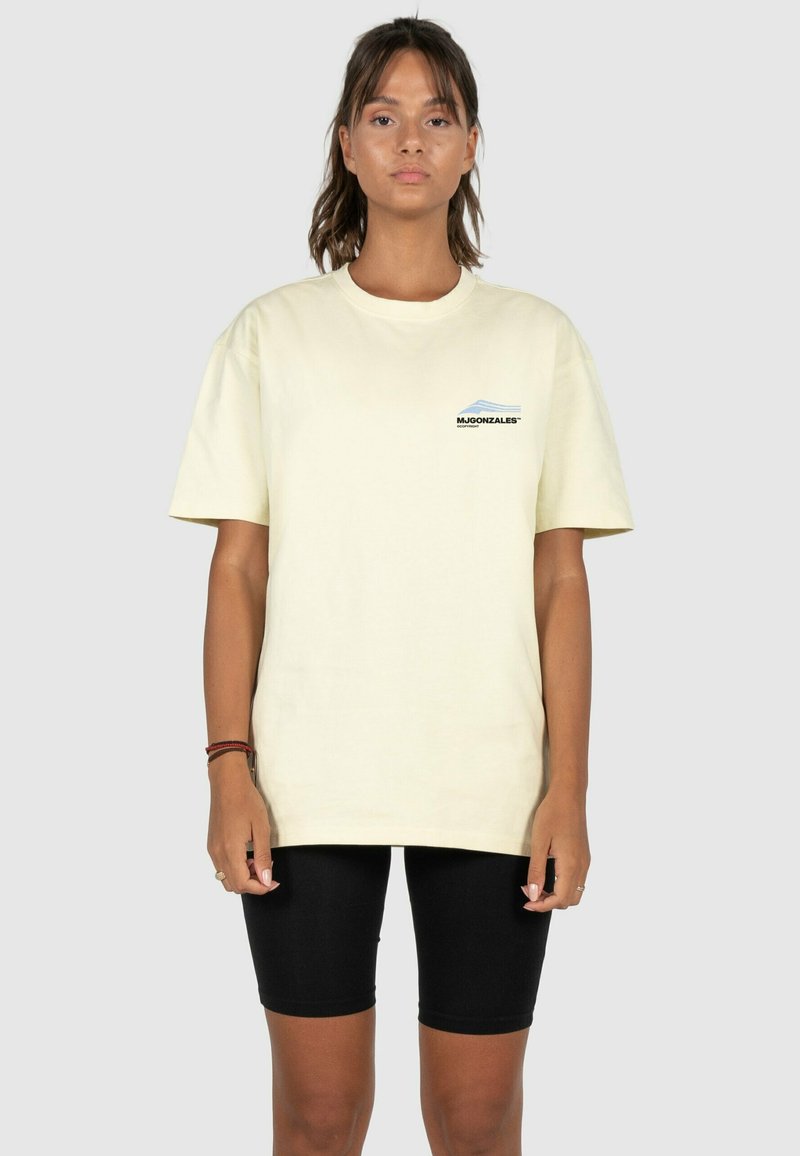 Light yellow cotton t-shirt with a crew neck, short sleeves, and a small blue and black logo on the left chest, paired with black shorts.