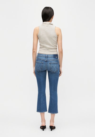 Γυναίκα που στέκεται με την πλάτη, φοράει αμάνικη ριγέ ζιβάγκο μπλούζα, κοντά τζιν παντελόνια και μαύρα ψηλοτάκουνα σανδάλια.