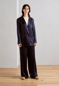 Blazer albastru marin cu două rânduri de nasturi și două buzunare frontale, asortat cu pantaloni largi. Șlapi galbeni cu vârful pătrat, textură de țesătură netedă.