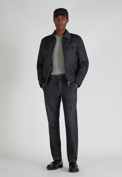 Giacca bomber nera, maglione grigio a costine, pantaloni in denim scuro e scarpe nere. Il modello si trova su uno sfondo grigio chiaro.