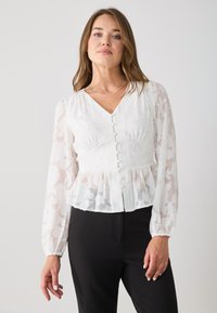 White peplum blouse with sheer, patterned long sleeves, v-neckline, and center button closure. Features subtle floral texture and ruffles.