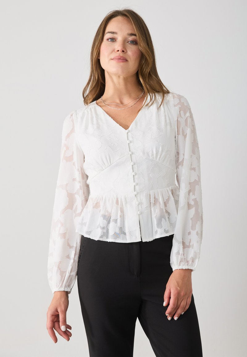 White peplum blouse with sheer, patterned long sleeves, v-neckline, and center button closure. Features subtle floral texture and ruffles.
