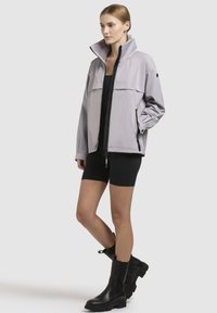 Light gray waterproof jacket with high collar, front pocket, and black zippers. Worn over a black top and shorts, paired with black ankle boots.