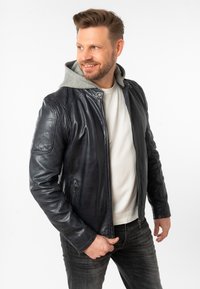 Marineblaue Lederjacke mit grauem Kapuzenfutter, Reißverschluss, Seitentaschen und strukturierten Schulterakzenten. Über einem weißen Hemd getragen.