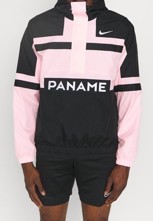 Mężczyzna ubrany w czarną i różową wiatrówkę Nike z napisem „PANAME”, w połączeniu z czarnymi szortami Nike na jednolitym tle.