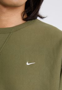 Zbližnji posnetek vratu in rame osebe, oblečene v olivno zeleno pulover s belim logotipom Nike na levi strani prsnega koša.