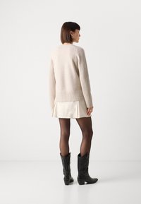 Beige strikkegenser med ribbestruktur, sammen med en kremfarget plisserte skjørt, gjennomsiktige svarte strømpebukser, og svarte cowboystøvler med detaljer.