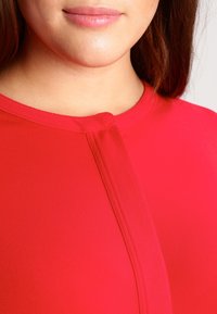 Röd blus med rund halsringning och en liten veck fram. Tillverkad av mjukt tyg, har subtila sömmar längs kanterna.