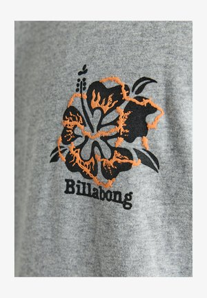 Grijze stoffen t-shirt met een zwart en oranje hibiscus bloemenontwerp met daaronder het woord "Billabong" in een vet lettertype.
