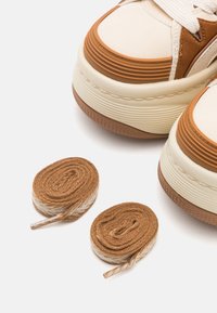Le sneakers bicolore presentano una parte superiore color crema con dettagli marroni, punta rotonda, suola testurizzata e due paia di lacci marroni per la personalizzazione.