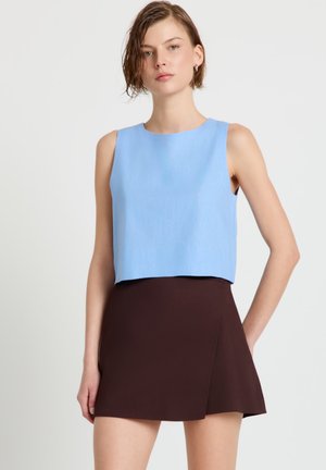 Frau mit kurzen Haaren trägt ein ärmelloses hellblaues Oberteil und einen dunkelbraunen asymmetrischen Minirock, steht vor einem einfarbigen Hintergrund.