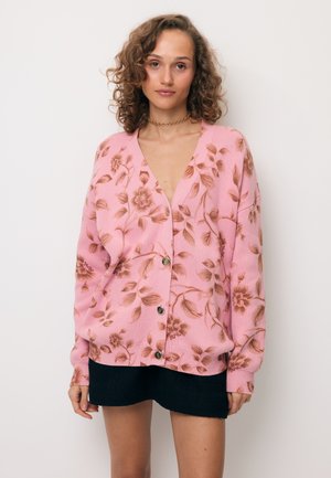 Vrouw met krullend haar draagt een roze gebloemde vest met knopen over een zwarte rok, staand tegen een effen lichte achtergrond.