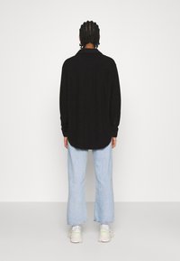 Chemise noire à manches longues avec un col, portée sur un pantalon en jean large bleu clair. Les caractéristiques notables incluent la texture lisse de la chemise et sa coupe décontractée.