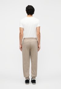 Beige joggingbroek met elastische tailleband en boorden, gecombineerd met een effen wit T-shirt, gezien van achteren. Zwarte sneakers maken de outfit compleet.