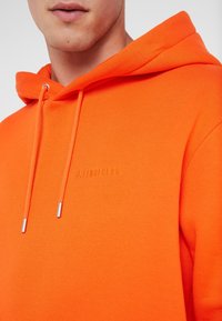 Orange hoodie gjord av strukturerad tyg, med en dragsko huva och präglad logo i matchande färg. Silverfärgade metalldelar på dragskorna.