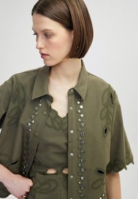 Veste vert olive avec broderies florales et clous métalliques décoratifs. Superposée sur un top vert assorti avec des accents en dentelle. Manches courtes.