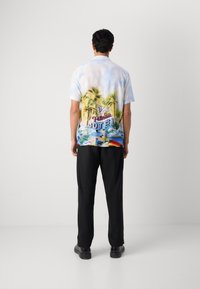 Camisa de manga corta, azul claro, con un colorido estampado tropical de palmeras y un gráfico de un motel; combinada con pantalones y zapatos negros.