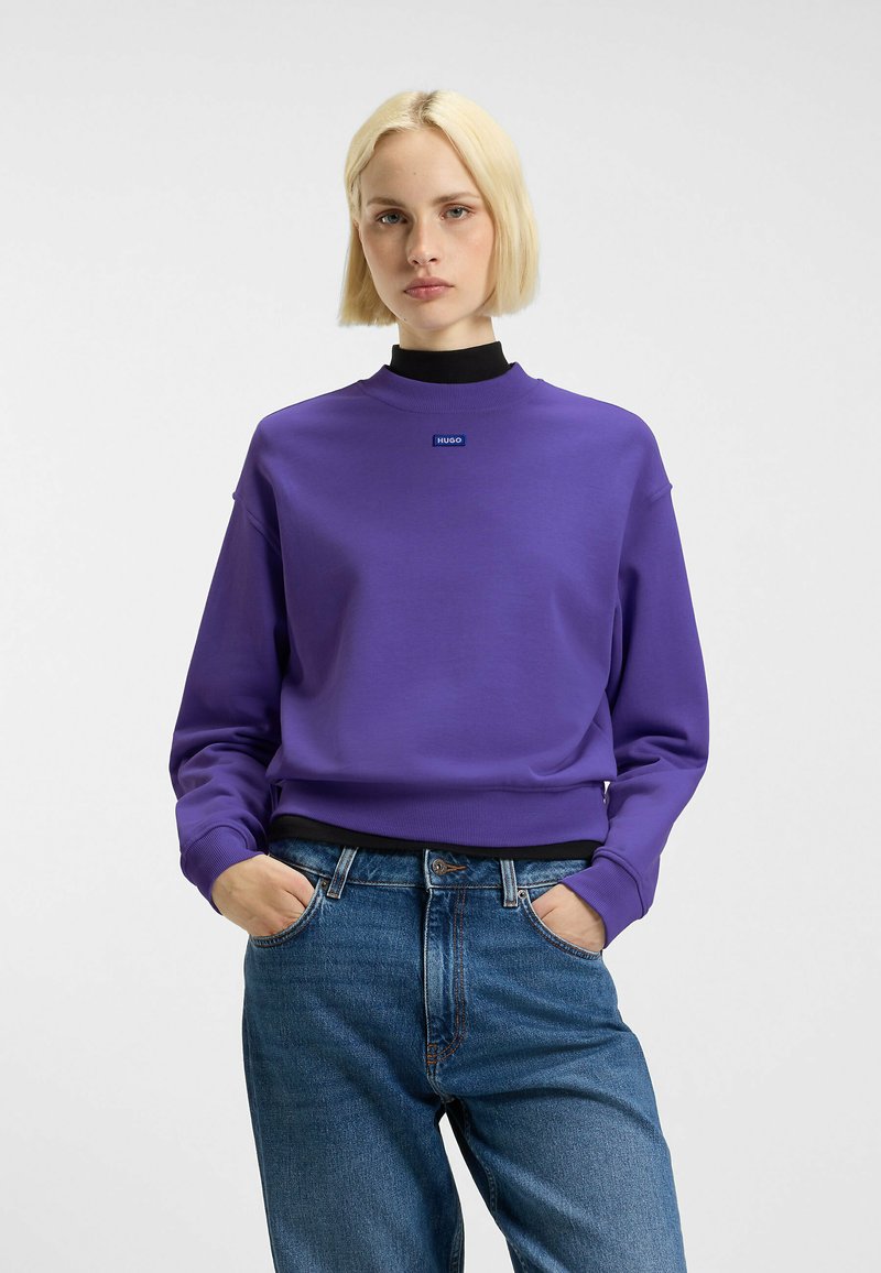 Paarse sweatshirt met een ronde hals en geribbelde manchetten, voorzien van een klein logo op de borst, gecombineerd met een blauwe spijkerbroek.