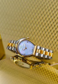 Kultainen ja hopeanvärinen kello, jossa on pisaran muotoinen kuori, sininen helmitaulu ja metallinen ranneke, jossa on vuorotellen kultaisia ja hopeanvärisiä linkkejä.