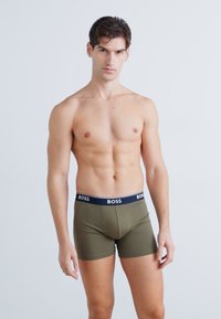 Boxeri de culoare verde măslin, cu un elastic negru etichetat "BOSS". Materialul pare să fie neted și elastic, evidențiind o siluetă fit.